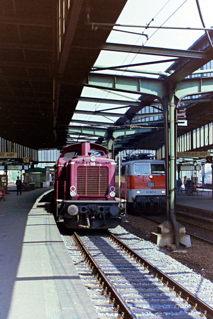 Eine rote Diesellok (211 084) steht in Duisburg Hbf an Gleis 6 zur Fahrt nach Kleve.