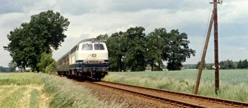 Ein Zug durchfährt bei Xanten eine Landschaft mit Feldern und Bäumen im Hintergrund. Lok ist 215 021